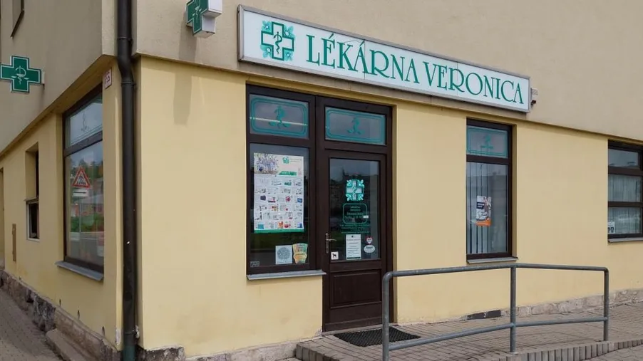 Vstup do lékárny Veronika s bezbariérovým přístupem a vývěskou na oknech, umístěné v přízemí žluté budovy na rohu.